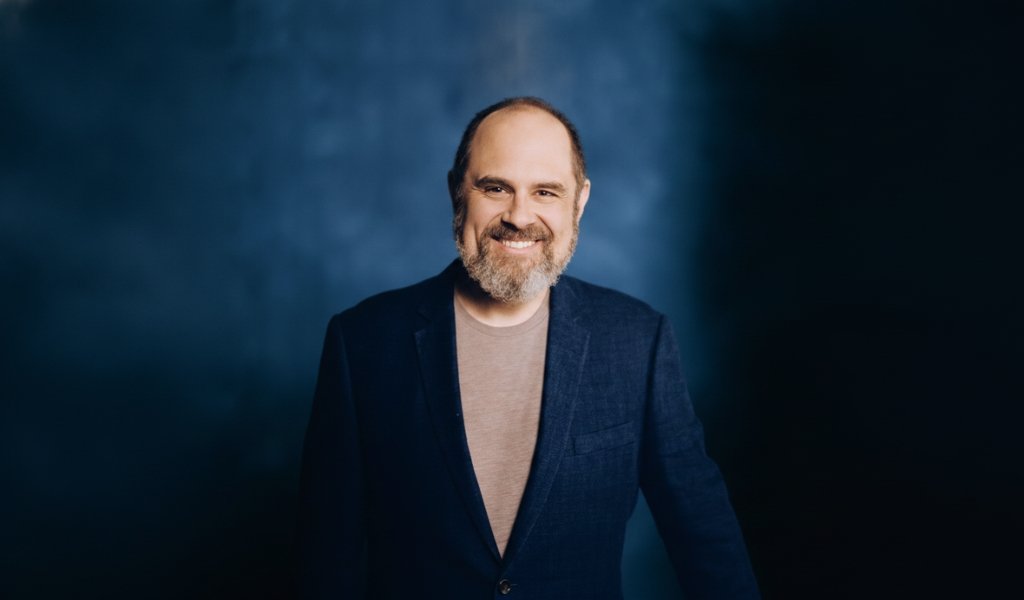 Craig Mazin smiling in a blue suit blazer against a textured blue background