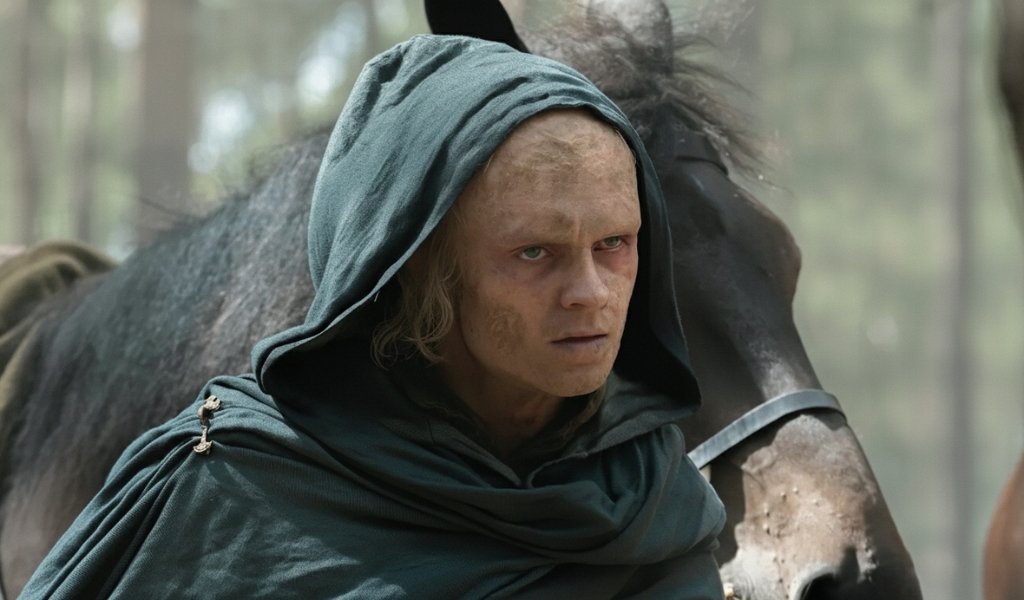 Tom Glynn-Carney as a scarred and wounded Aegon II Targaryen wearing a green hood with a horse in the background during his exile in House of the Dragon Season 3.